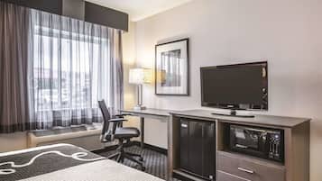 A hotel room with a bed, a desk with a television, a refrigerator, and a chair.