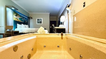 Habitación de hotel con una bañera grande, cama con ropa de cama blanca, un cuadro en la pared y un escritorio con lámpara.