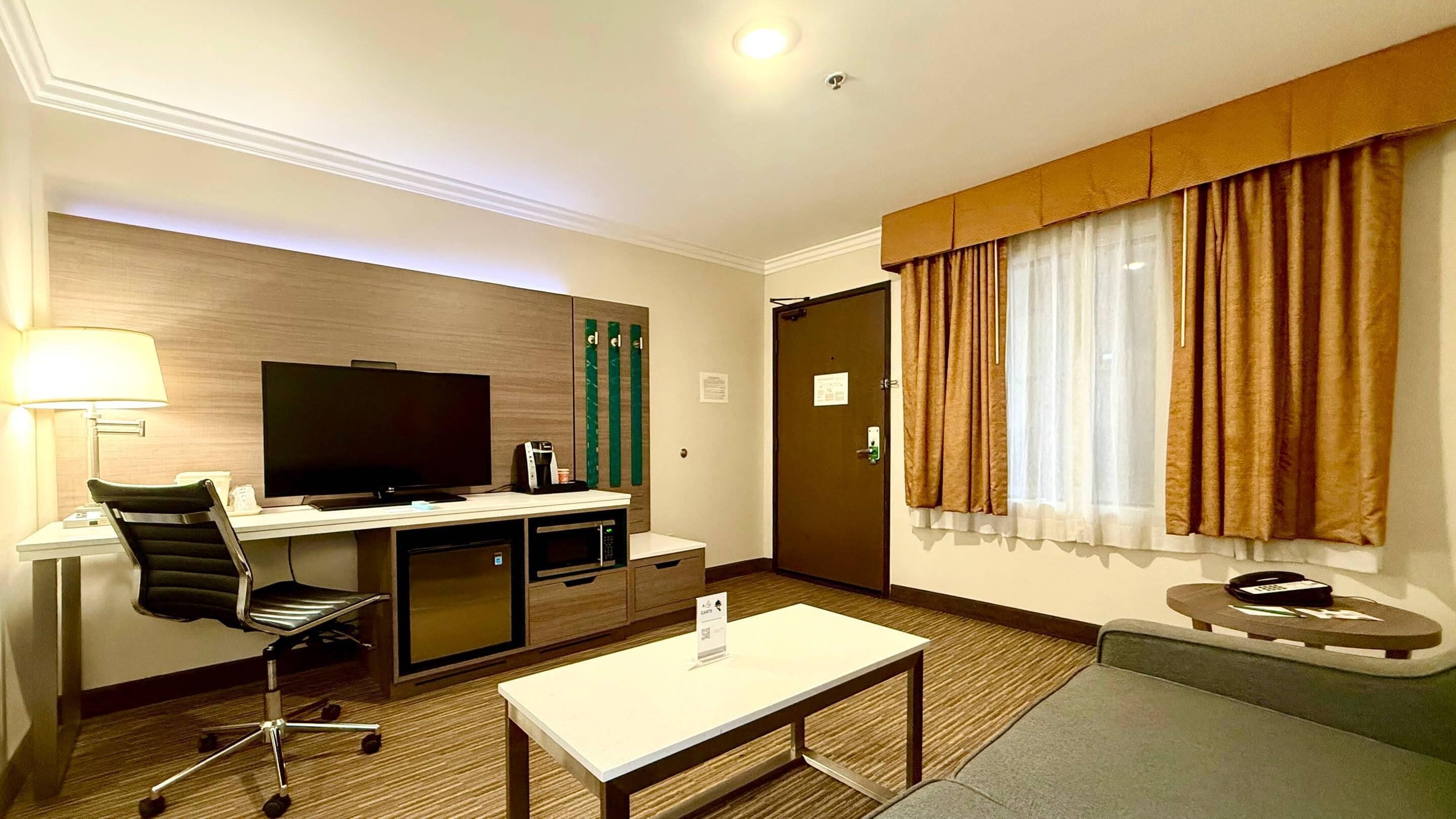 A hotel room with a flat-screen TV, a desk with a chair, a coffee machine, a mini-fridge, a small table, a sofa, and a window with curtains.
