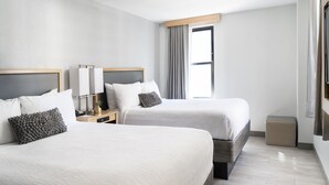 A hotel room with two beds, a window with curtains, a bedside table with a lamp, and a small ottoman.