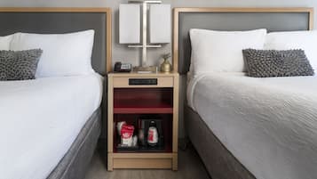 Une chambre d’hôtel avec deux lits, chacun doté de draps blancs et d’oreillers gris. On y trouve une table de chevet avec un téléphone, une petite plante et une machine à café. La chambre présente un design moderne, avec une tête de lit en bois et un mur gris.