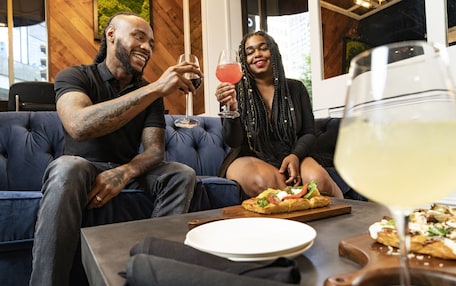 A couple sitting on a blue couch with a table in front of them, holding drinks and a pizza.