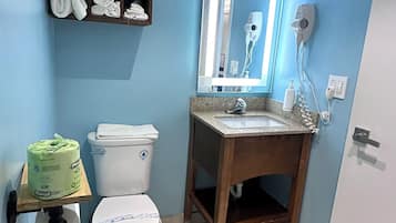 A bathroom with a toilet, sink, and mirror. There is a shelf with towels and a trash can.