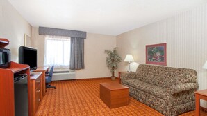 A hotel room with a patterned carpet, a sofa, a desk with a computer, a television, a lamp, and a window with curtains.