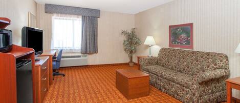Una habitación de hotel con alfombra estampada, un sofá, un escritorio con computadora, un televisor, una lámpara y una ventana con cortinas.