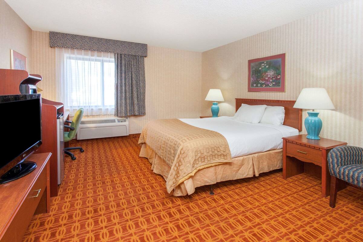 Une chambre d’hôtel comprenant un lit, deux tables de chevet avec des lampes, un bureau avec une télévision, une chaise et une fenêtre avec des rideaux.