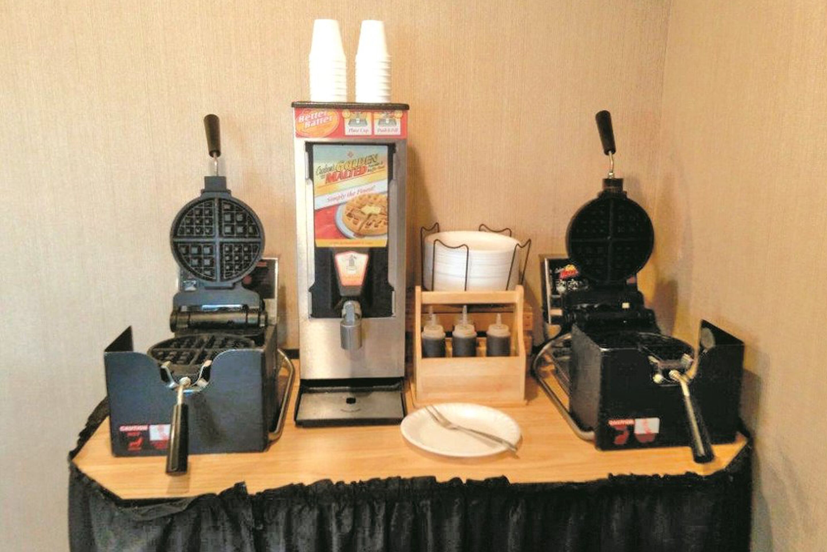 A breakfast bar with waffle makers, a waffle machine, a waffle dispenser, and condiments.