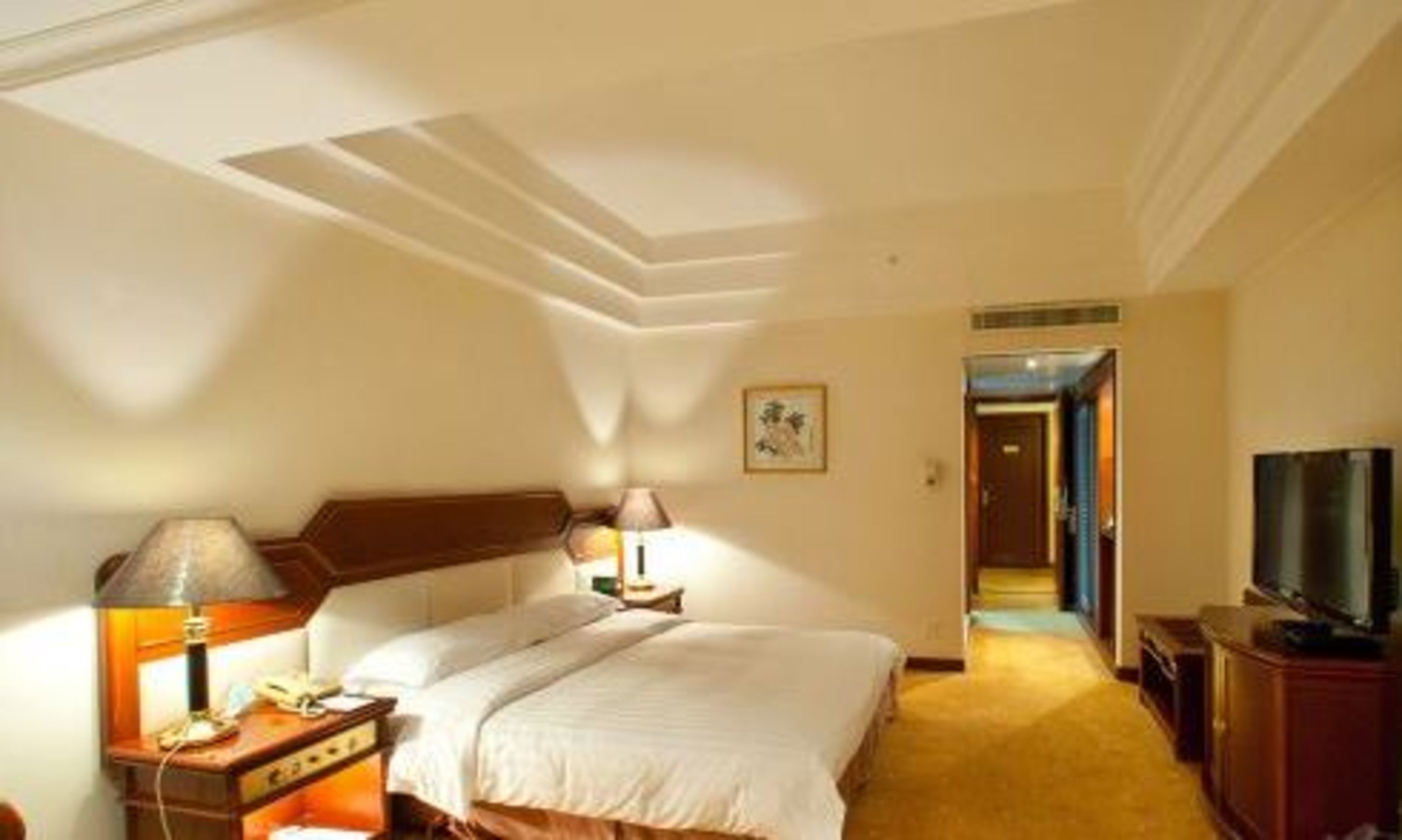 Una habitación de hotel con cama, mesita de noche, lámpara, teléfono, televisión y un cuadro en la pared.