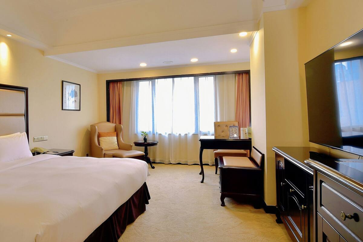Une chambre d’hôtel avec un grand lit, un bureau, une chaise, une télévision et une fenêtre avec des rideaux.