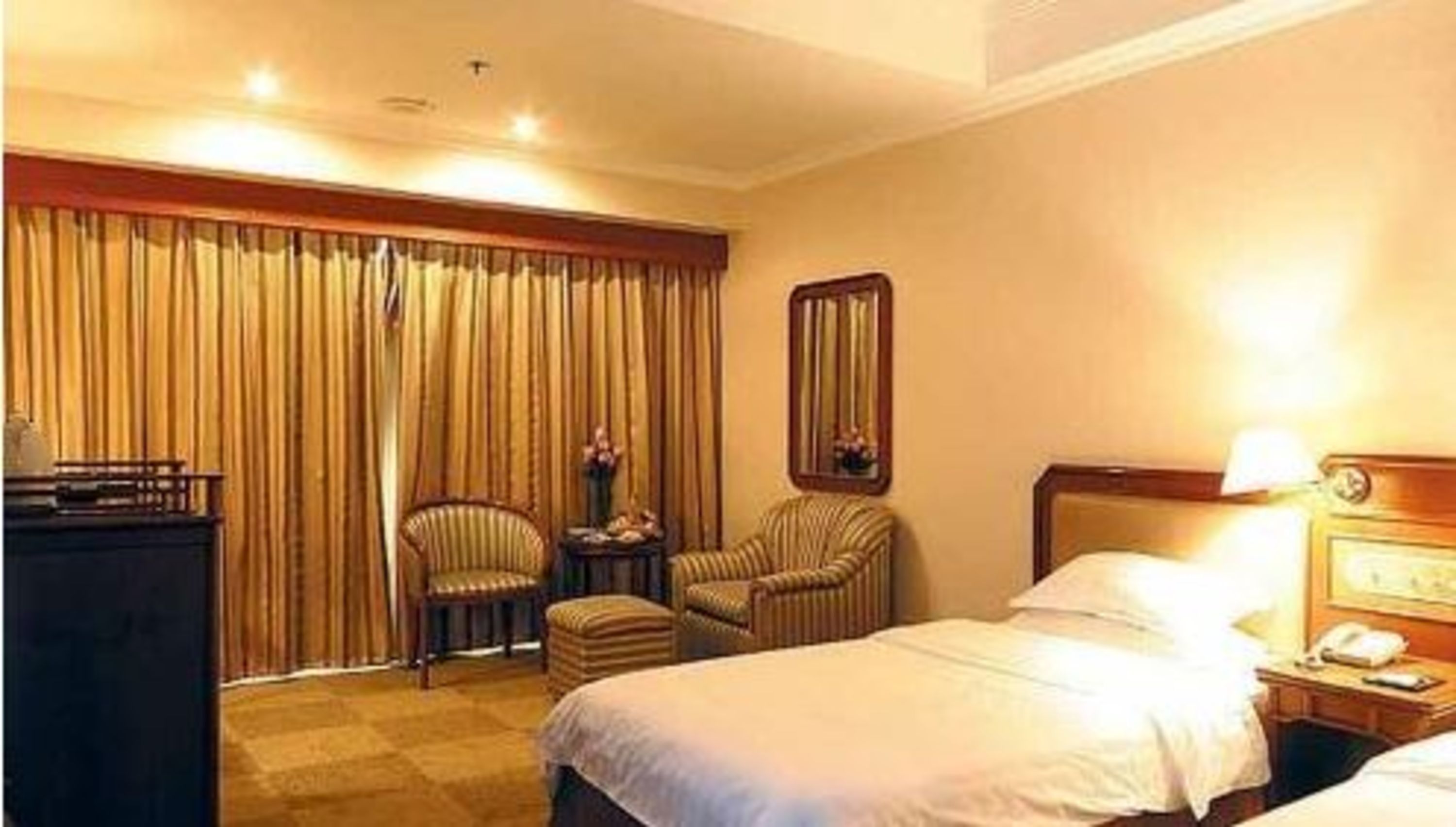 A hotel room with a bed, a desk with a lamp, a chair, a small table with flowers, and a large window with curtains.