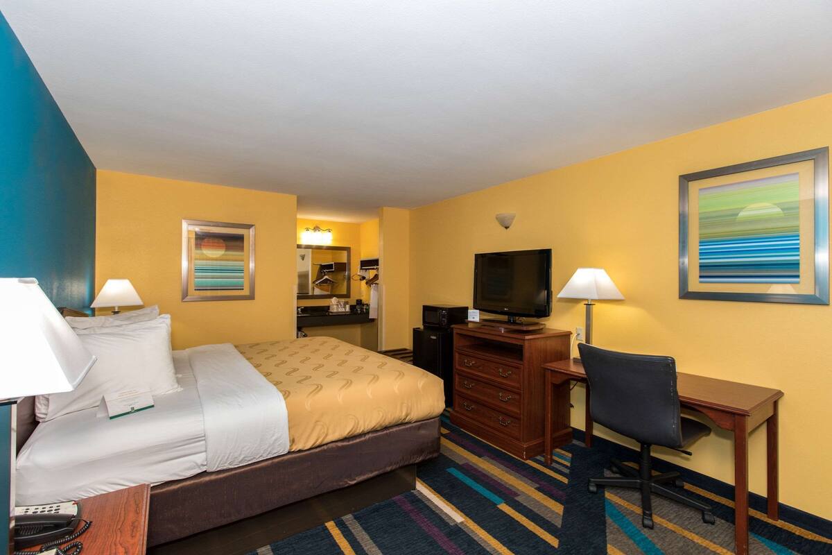 A hotel room with a bed, desk, chair, television, and a bathroom visible in the background.