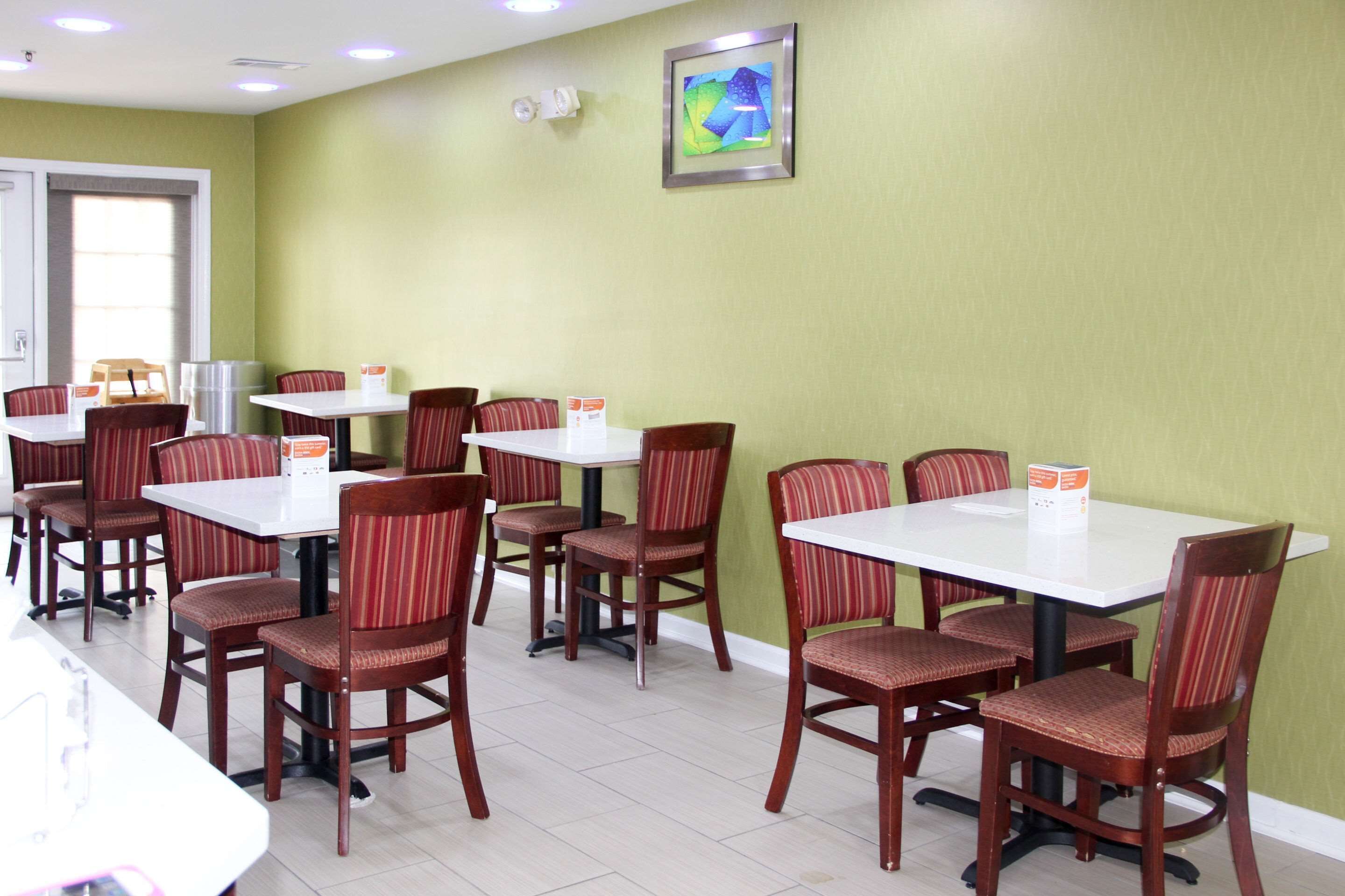 A dining area with multiple tables and chairs, a framed artwork on the wall, and a window with blinds.