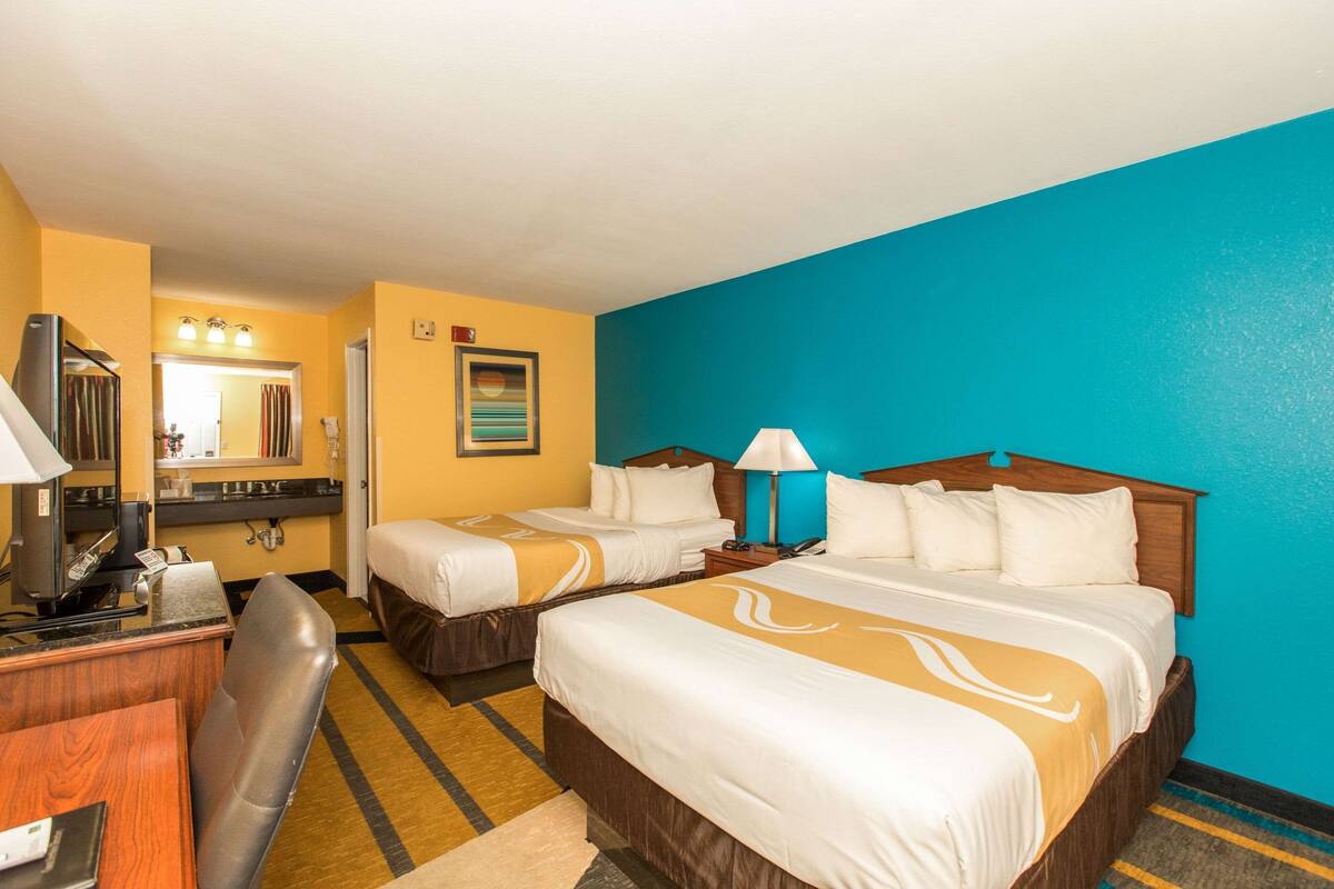 A hotel room with two beds, a desk, a chair, a desk lamp, a mirror, and a window.