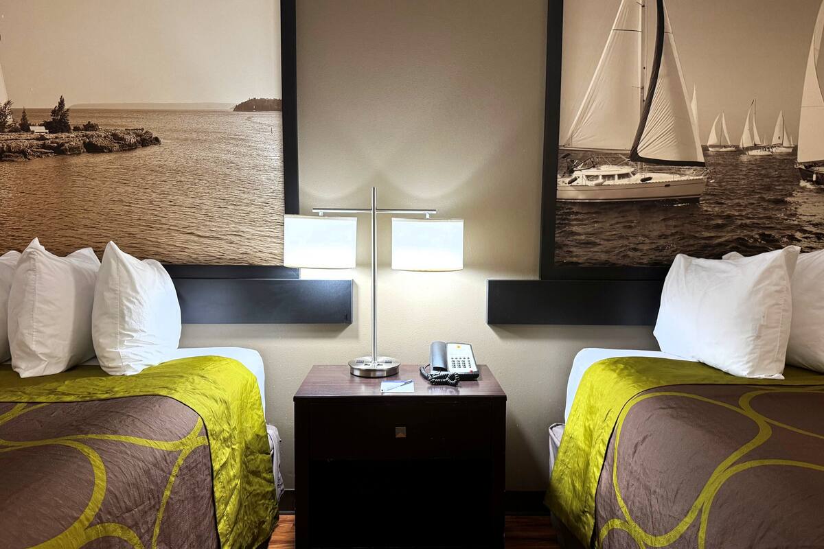 A hotel room with two beds, a nightstand with a lamp and phone, and a wall with a painting of sailboats.