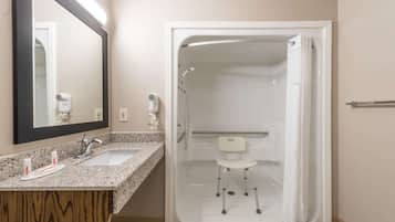 A bathroom with a granite countertop, a white sink, a mirror, a shower with a white chair, and a towel rack.
