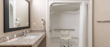 Une salle de bain avec un comptoir en granit, un lavabo blanc, un miroir, une douche avec une chaise blanche et un porte-serviettes.