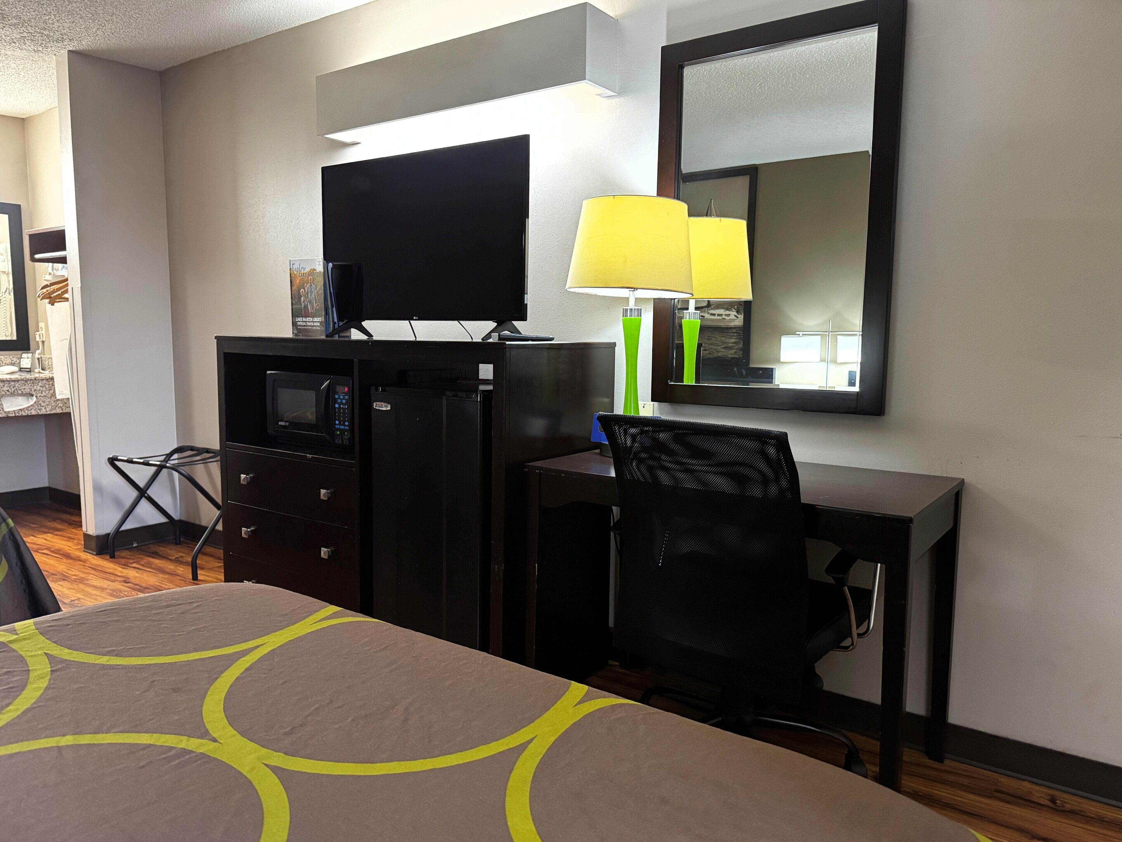 A hotel room with a bed, a desk with a chair, a television, a refrigerator, a microwave, a lamp, and a mirror.
