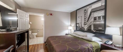 Habitación de hotel con cama, escritorio, televisor y baño visible a través de una puerta abierta.
