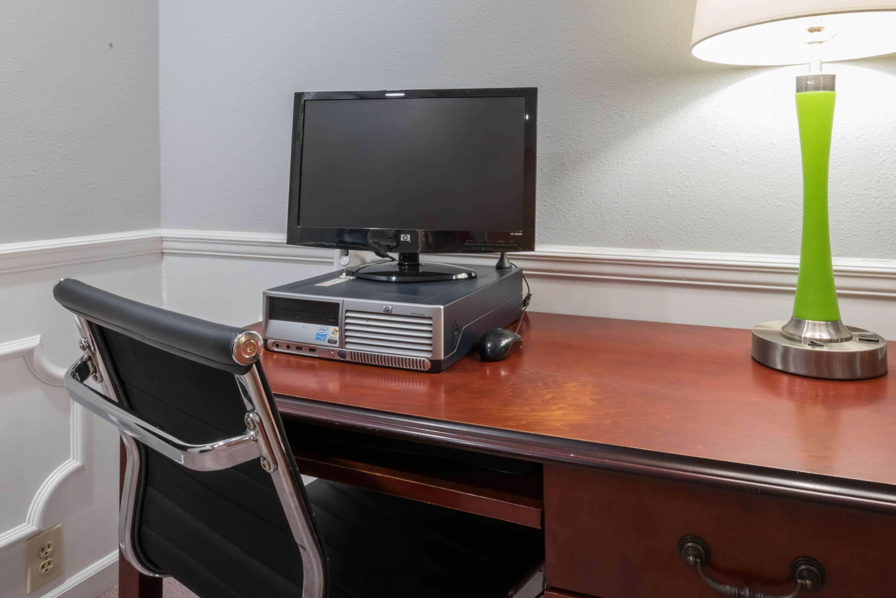Habitación de hotel con un escritorio de madera, una silla de oficina negra, un monitor de pantalla plana, una impresora y una lámpara de pie verde.