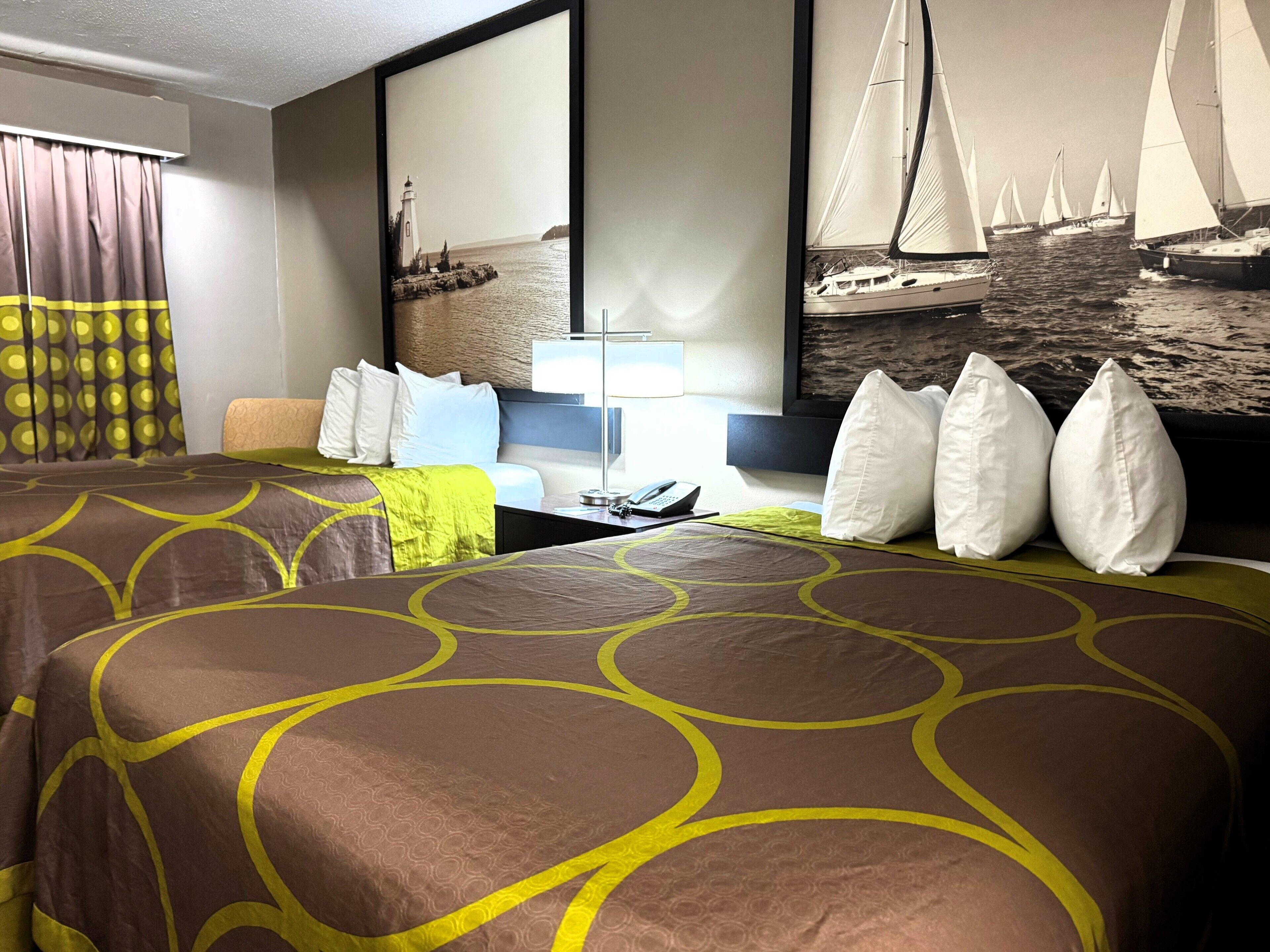 A hotel room with a bed, pillows, a desk with a phone, a lamp, and a painting of sailboats.