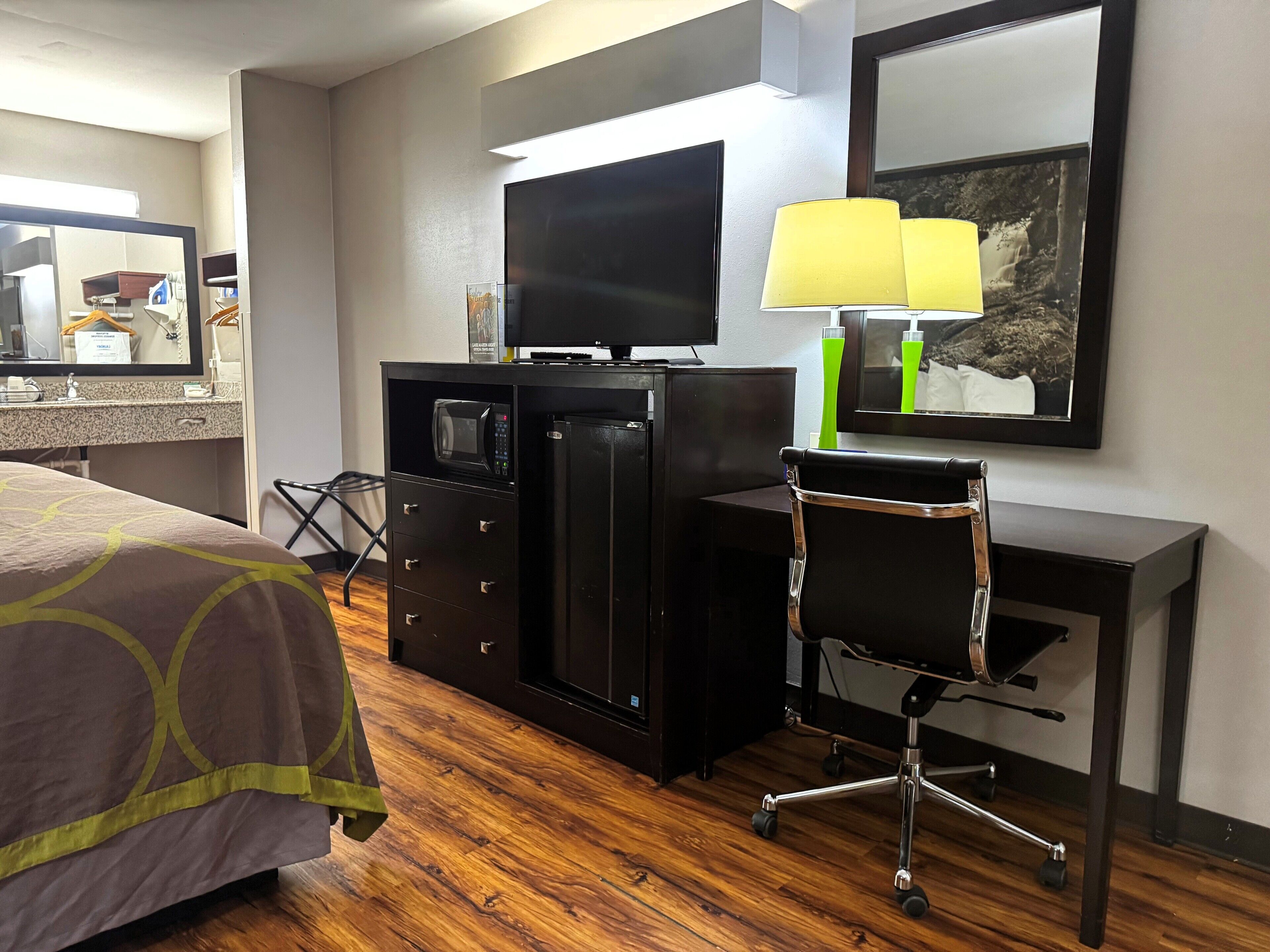 A hotel room with a bed, a desk with a flat-screen TV, a chair, a desk lamp, and a framed picture on the wall.