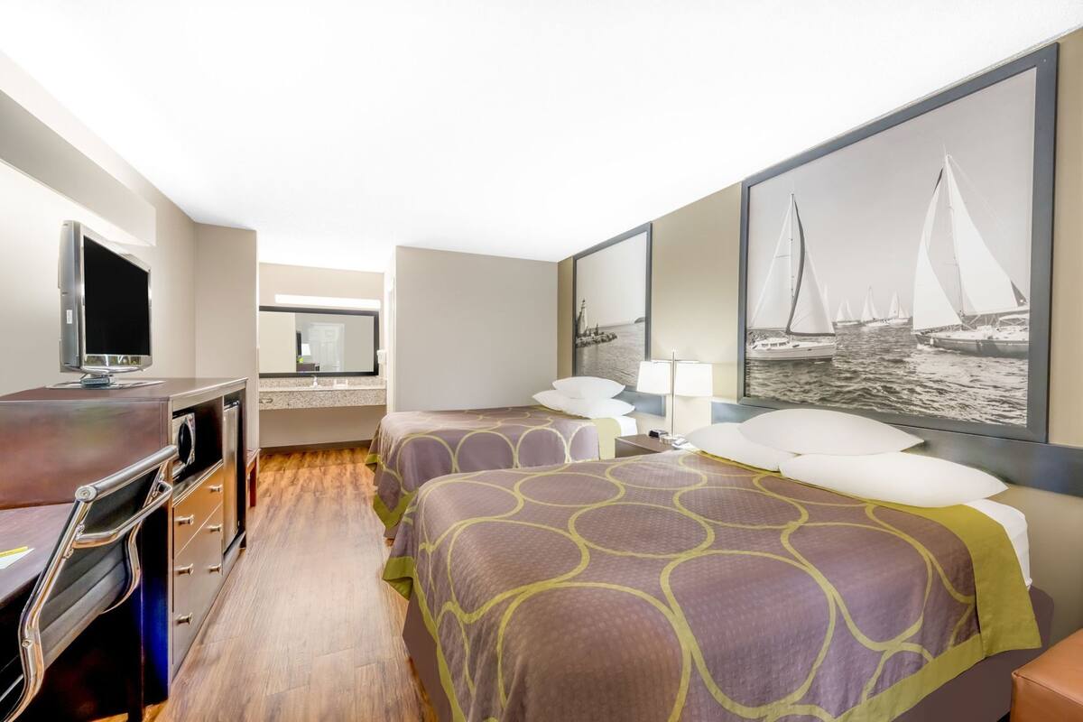 A hotel room with a TV, a desk, a bed with patterned bedding, a mirror, a window with a sailboat view, and a wall-mounted artwork.
