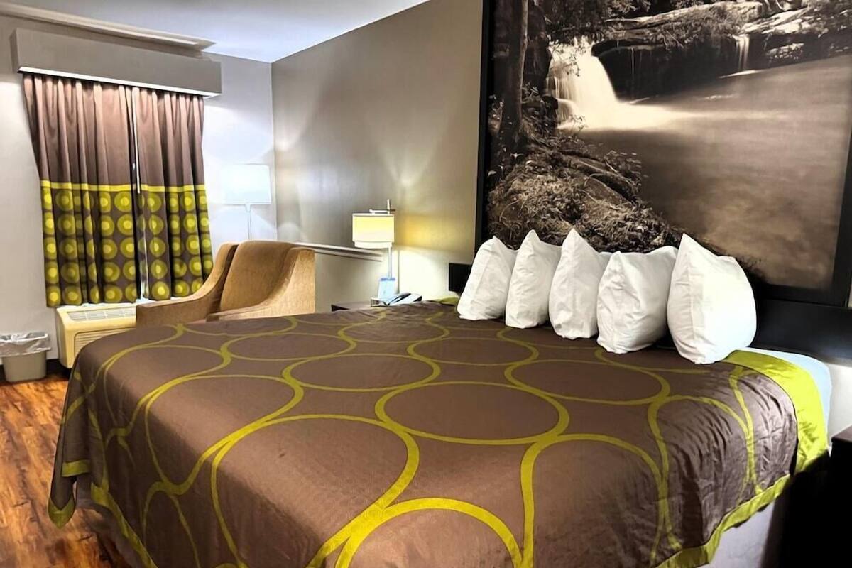 A hotel room with a large bed, a chair, a desk with a lamp, and a large wall painting.
