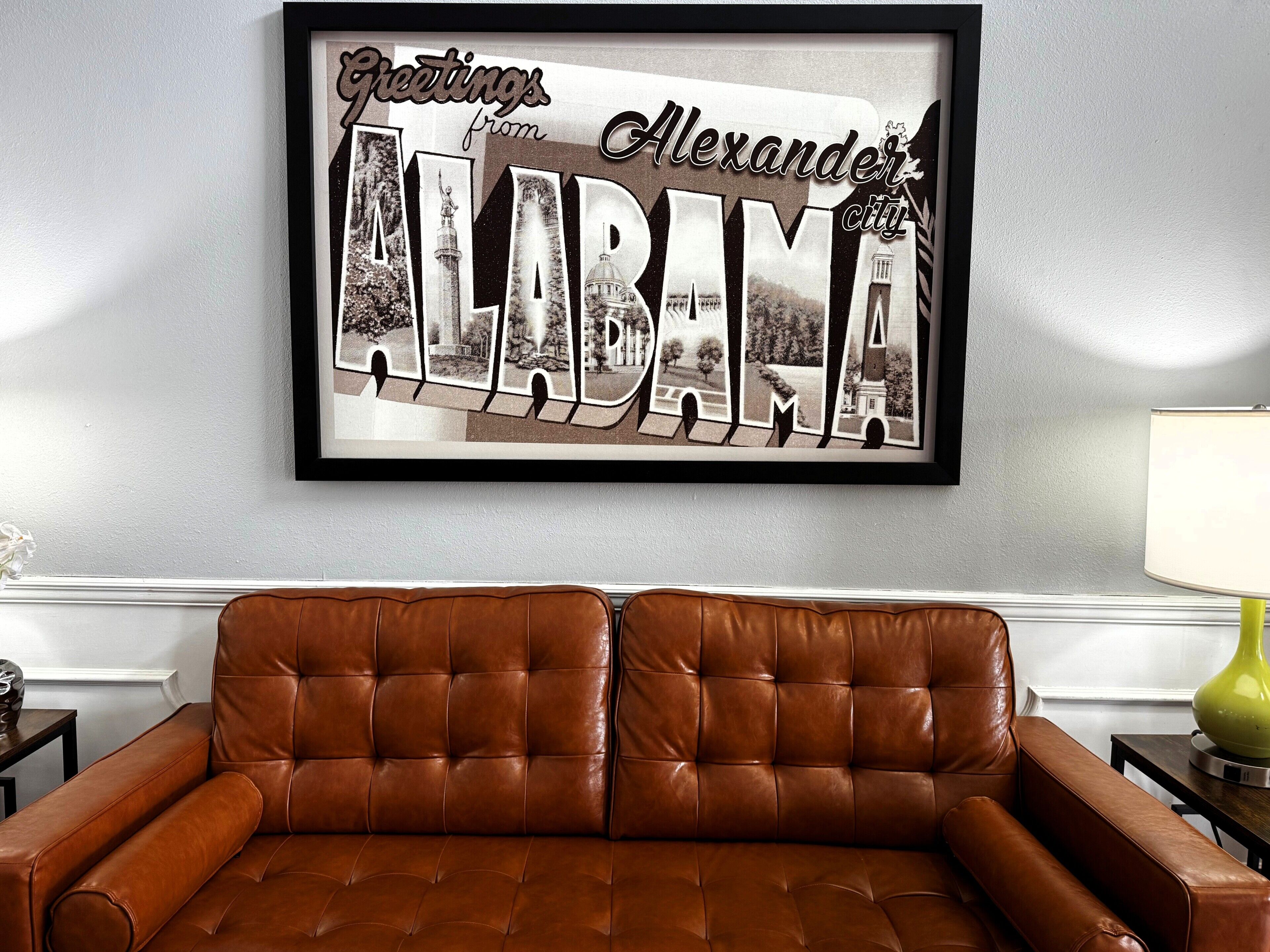 A framed 'Greetings from Alabama' sign with architectural photos, a brown leather sofa, and a lamp on a table.