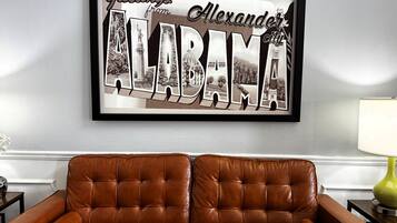 A framed 'Greetings from Alabama' sign with architectural photos, a brown leather sofa, and a lamp on a table.