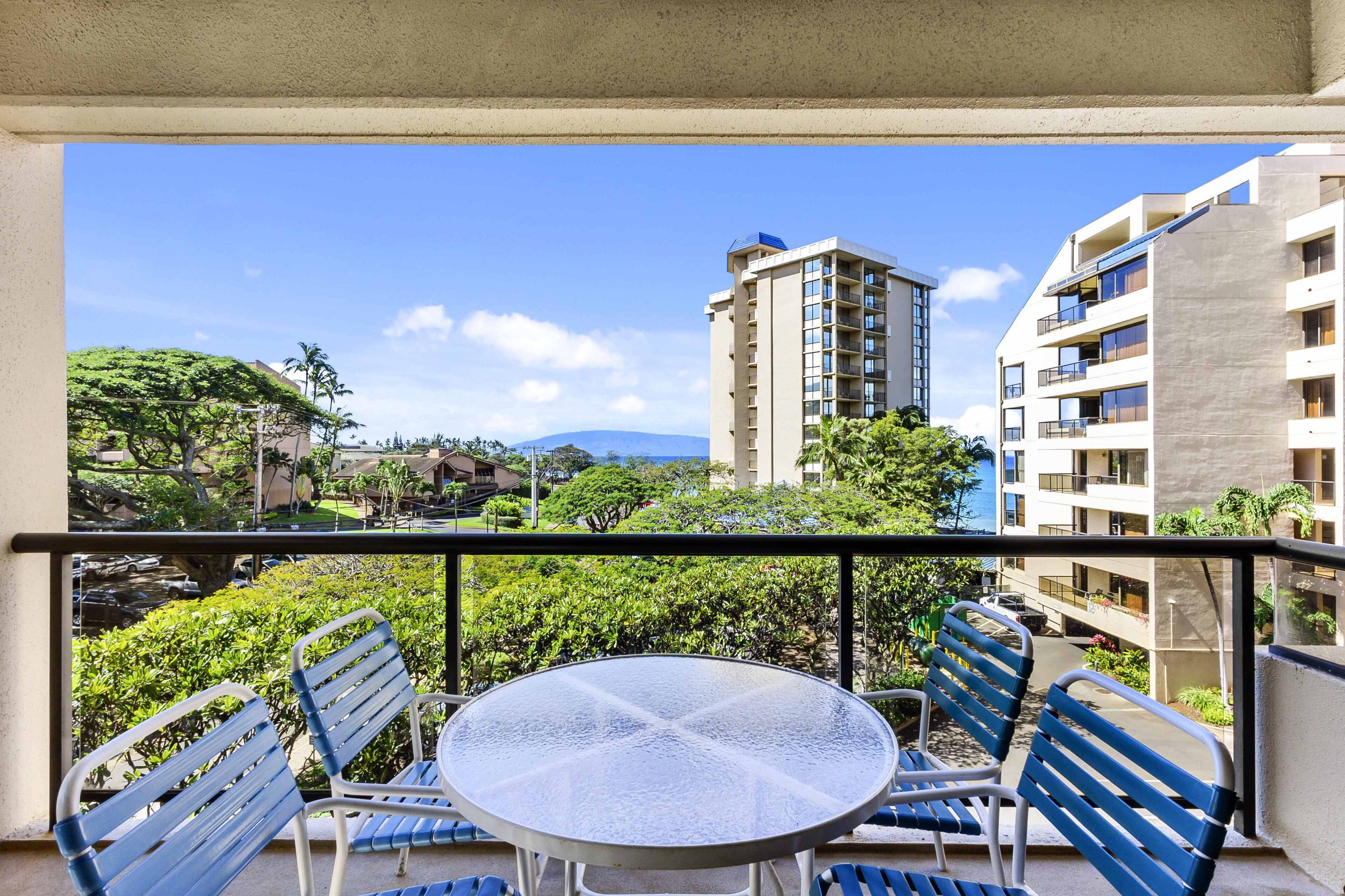 A balcony with a round table and blue chairs, overlooking a cityscape with buildings and greenery.