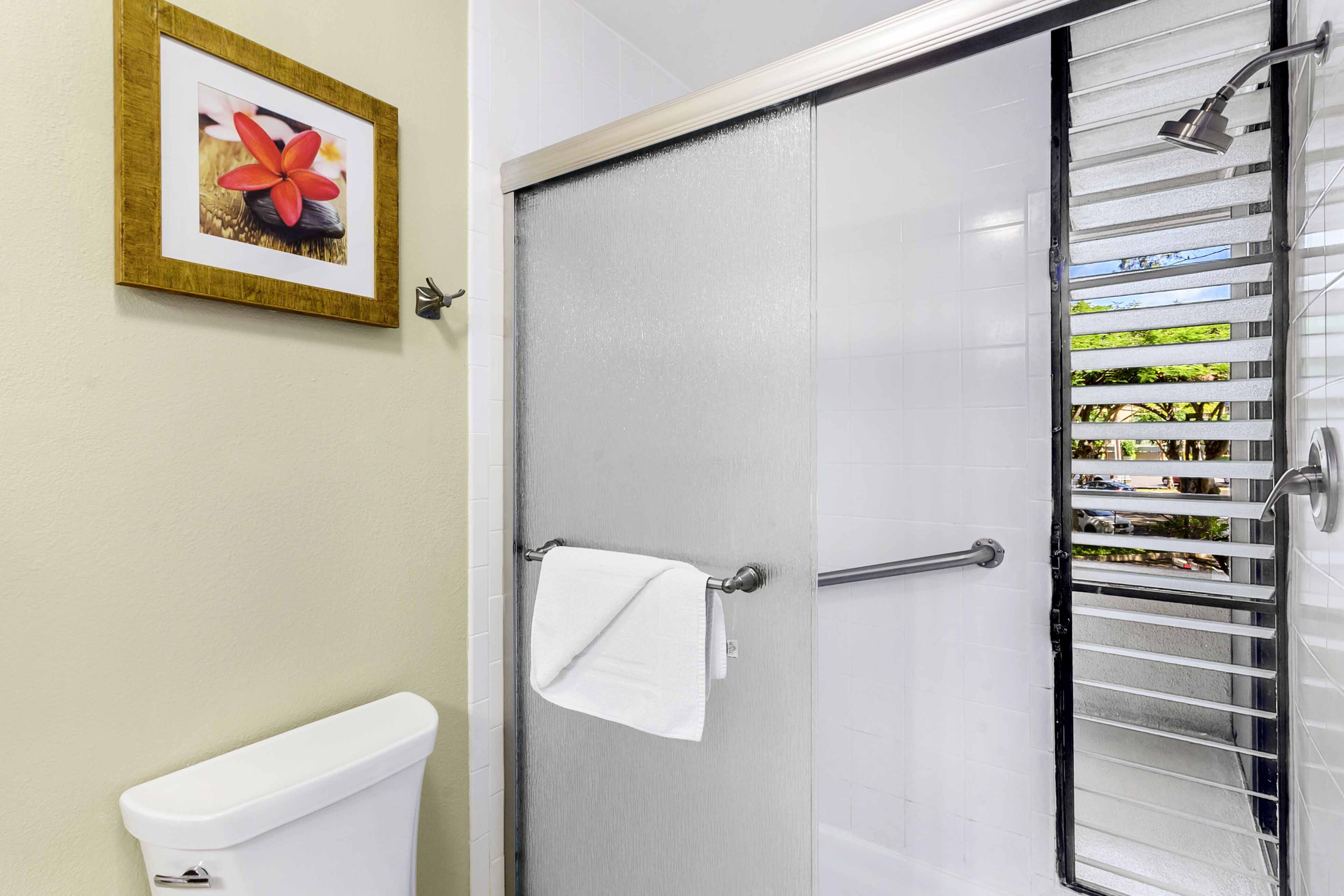 Un baño con inodoro, un perchero con toalla blanca, ducha con puerta de cristal y un cuadro en la pared.