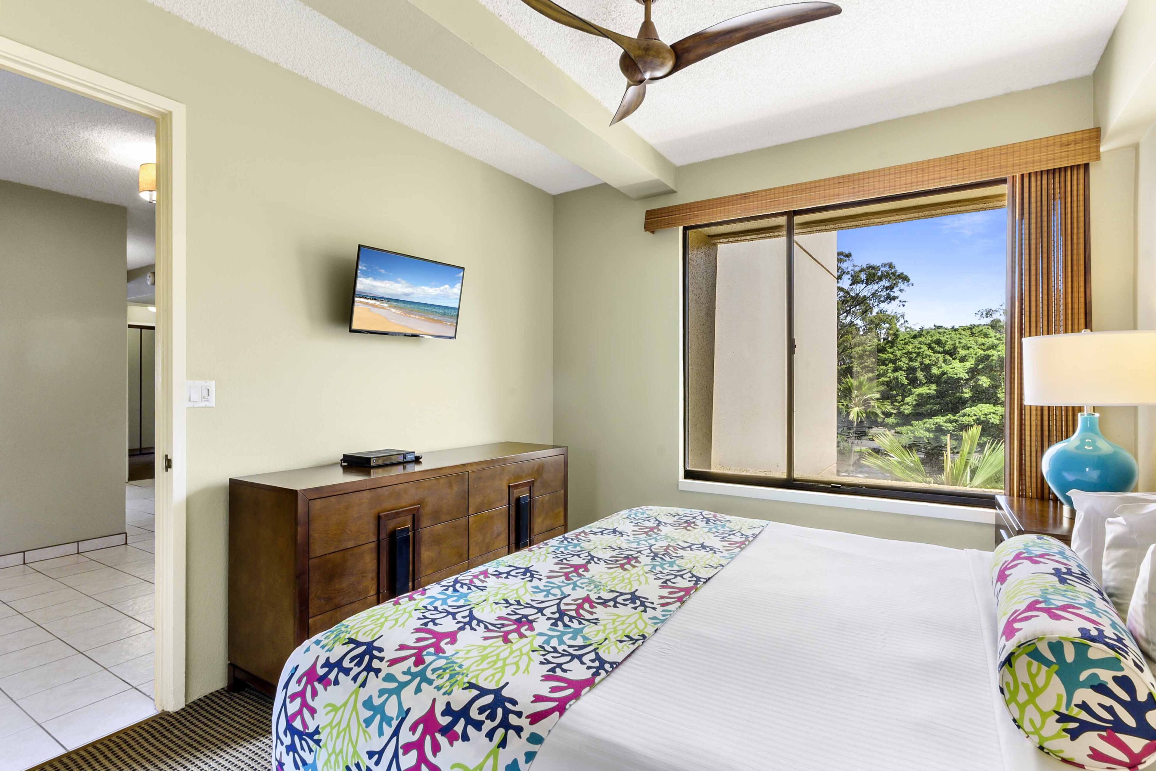 Habitación de hotel con cama, televisor montado en la pared, ventana con vistas a zonas verdes y ventilador de techo.