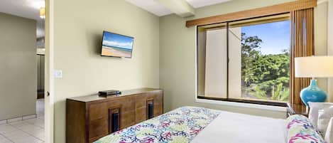 A hotel room with a bed, a TV mounted on the wall, a window with a view of greenery, and a ceiling fan.