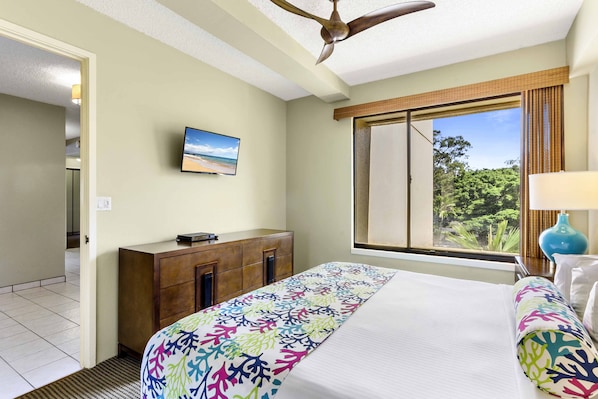 A hotel room with a bed, a TV mounted on the wall, a window with a view of greenery, and a ceiling fan. - Sands of Kahana (Lahaina)