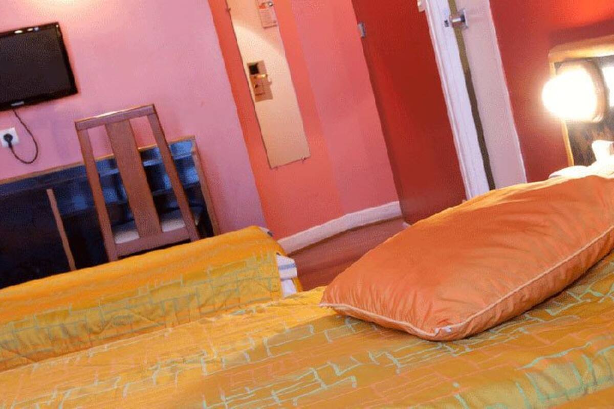 Un lit avec une literie orange et une tête de lit, un téléviseur fixé au mur et une chaise en bois.