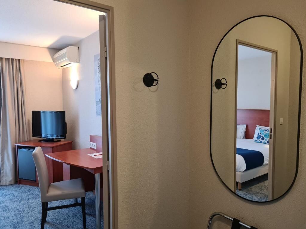 A hotel room with a bed, a desk with a television, a chair, a mirror, and a door leading to another room.