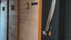 Een gang met een houten vloer, een zwarte deur met een zwaard erop en een lichtgekleurde muur met een structuur.