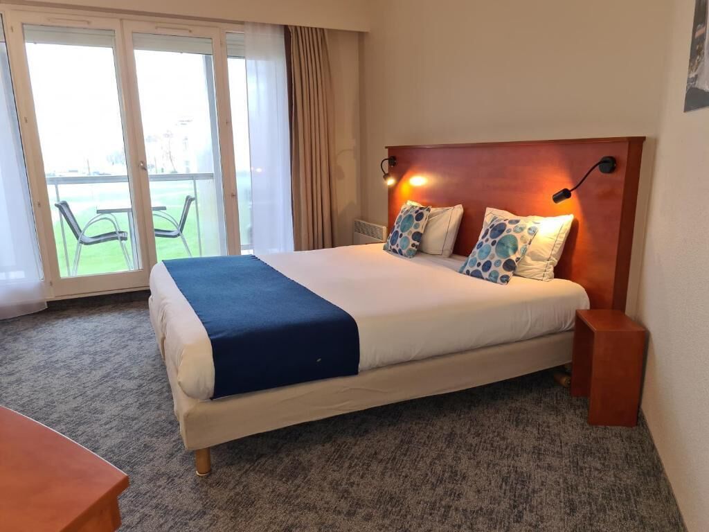 A hotel room with a bed, a desk, a chair, a window with curtains, and a view of the outdoors.