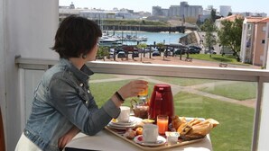 Een balkon met een ontbijttafel, een rode koffiezetapparaat en uitzicht op een jachthaven met boten en gebouwen.