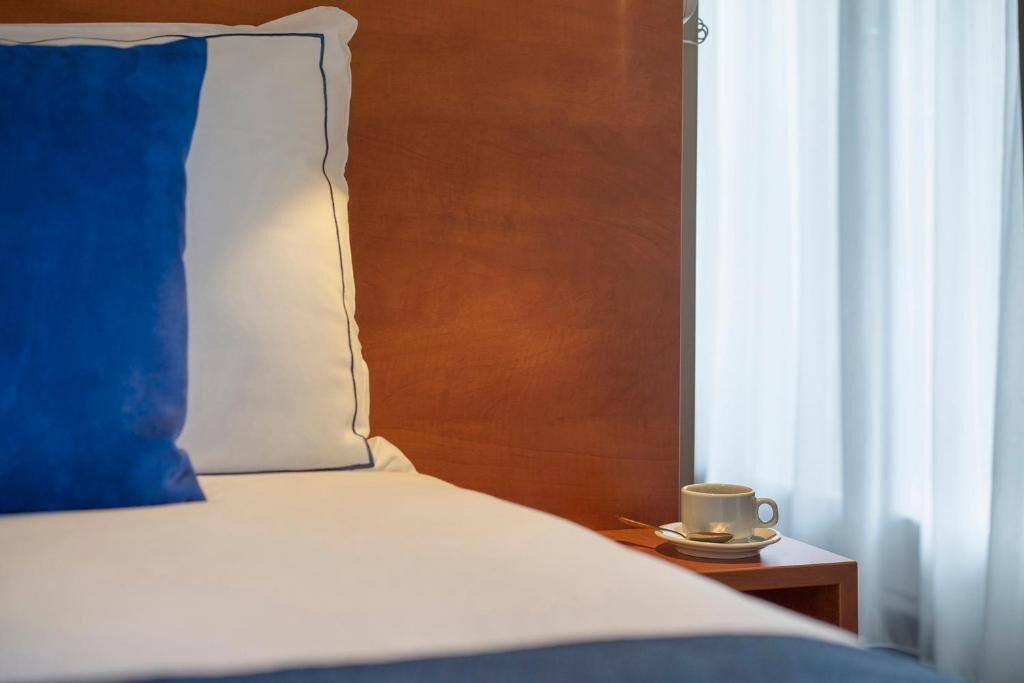 A neatly made bed with a blue and white pillow, a wooden headboard, and a bedside table with a cup and saucer.