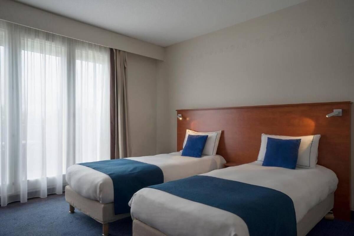 Une chambre d’hôtel avec deux lits, une tête de lit en bois et une grande fenêtre ornée de rideaux légers.