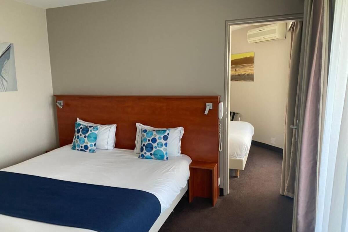 Une chambre d’hôtel avec un grand lit, deux oreillers, une tête de lit en bois, une table de chevet, un téléphone et une porte donnant sur une autre pièce.