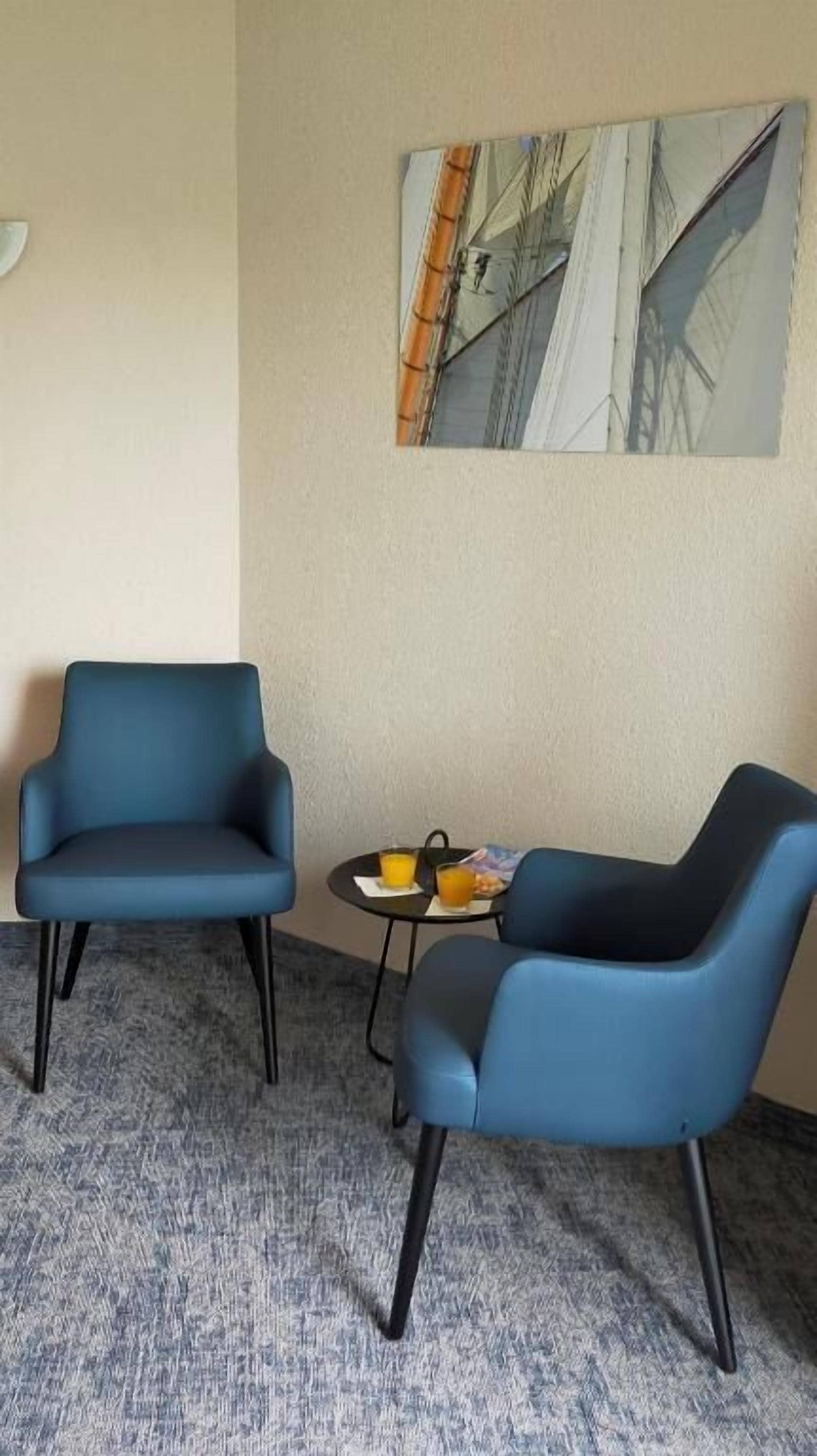 A room with two blue armchairs, a small table with a fruit plate, and a painting of a sailboat on the wall.