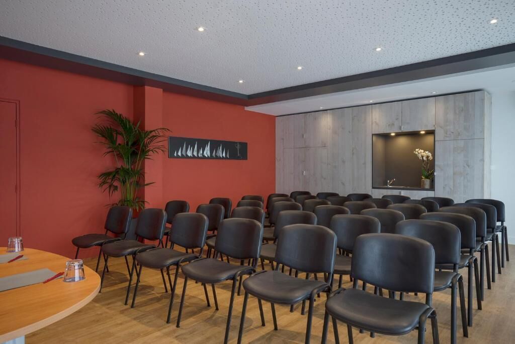 Une salle de conférence avec des rangées de chaises noires, une table en bois sur laquelle se trouve un verre, et une plante en pot.