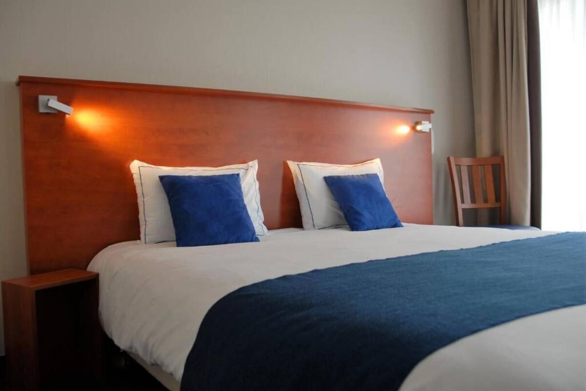 Une chambre d’hôtel avec une tête de lit en bois, deux oreillers, une couverture bleue et une table de chevet en bois.