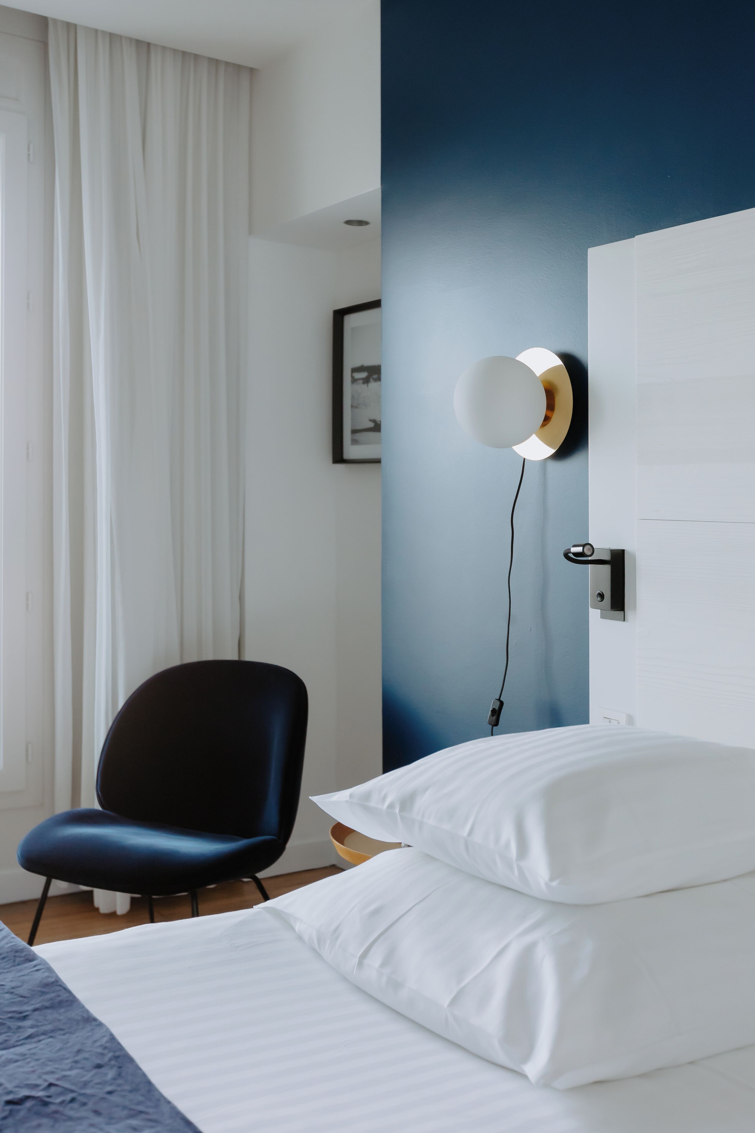 A hotel room with a bed, a chair, a wall-mounted light, and a door.