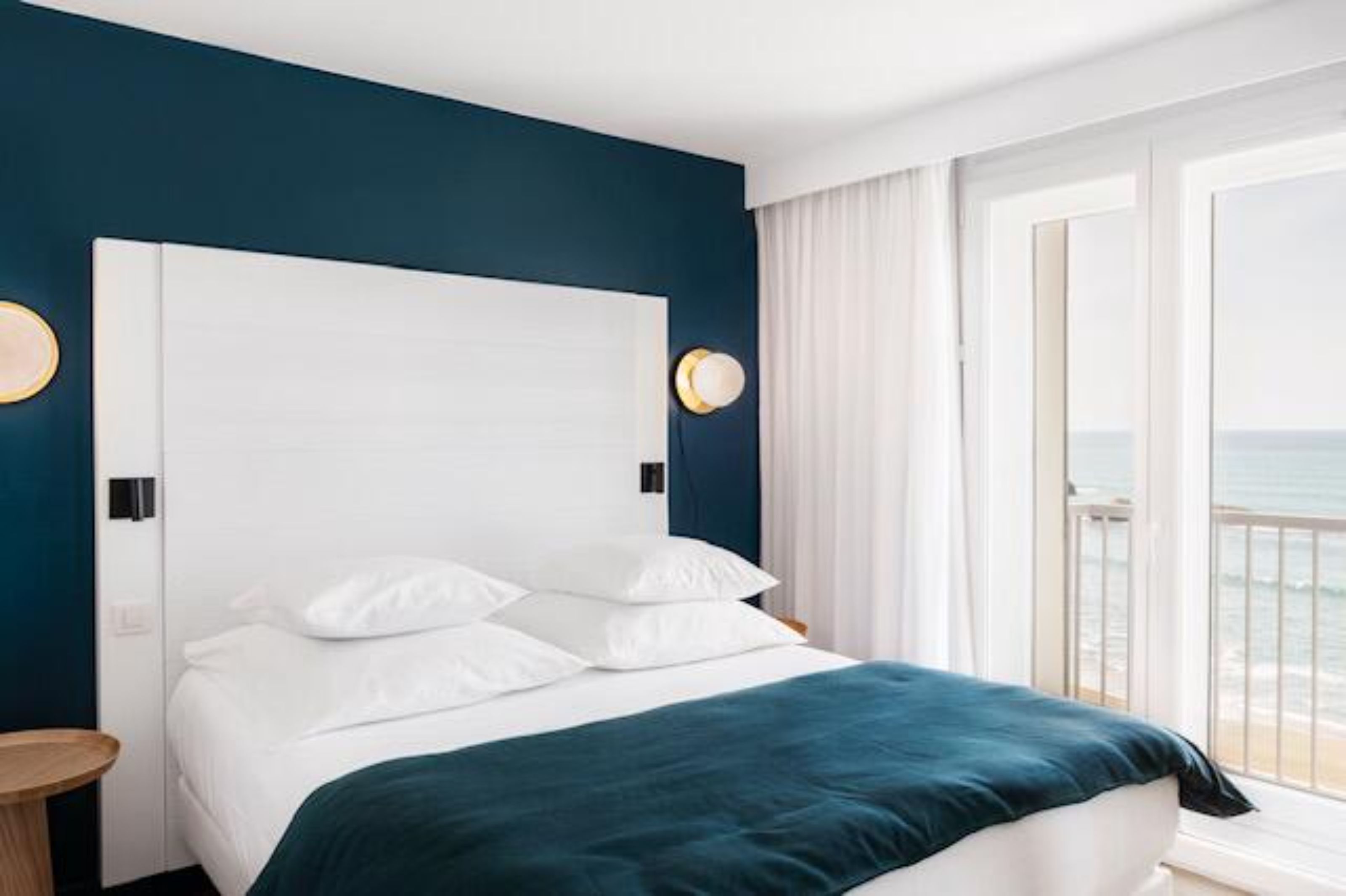 A hotel room with a large bed, white bedding, a teal throw blanket, and a view of the ocean through a balcony.
