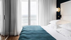 Une chambre à coucher avec un lit, des rideaux blancs, une vue sur la mer et une table de chevet avec une lampe.