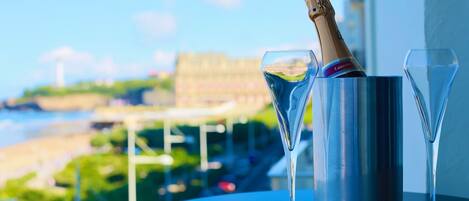 A champagne bottle in a metallic container with two glasses on a table, overlooking a scenic view of a beach and buildings.