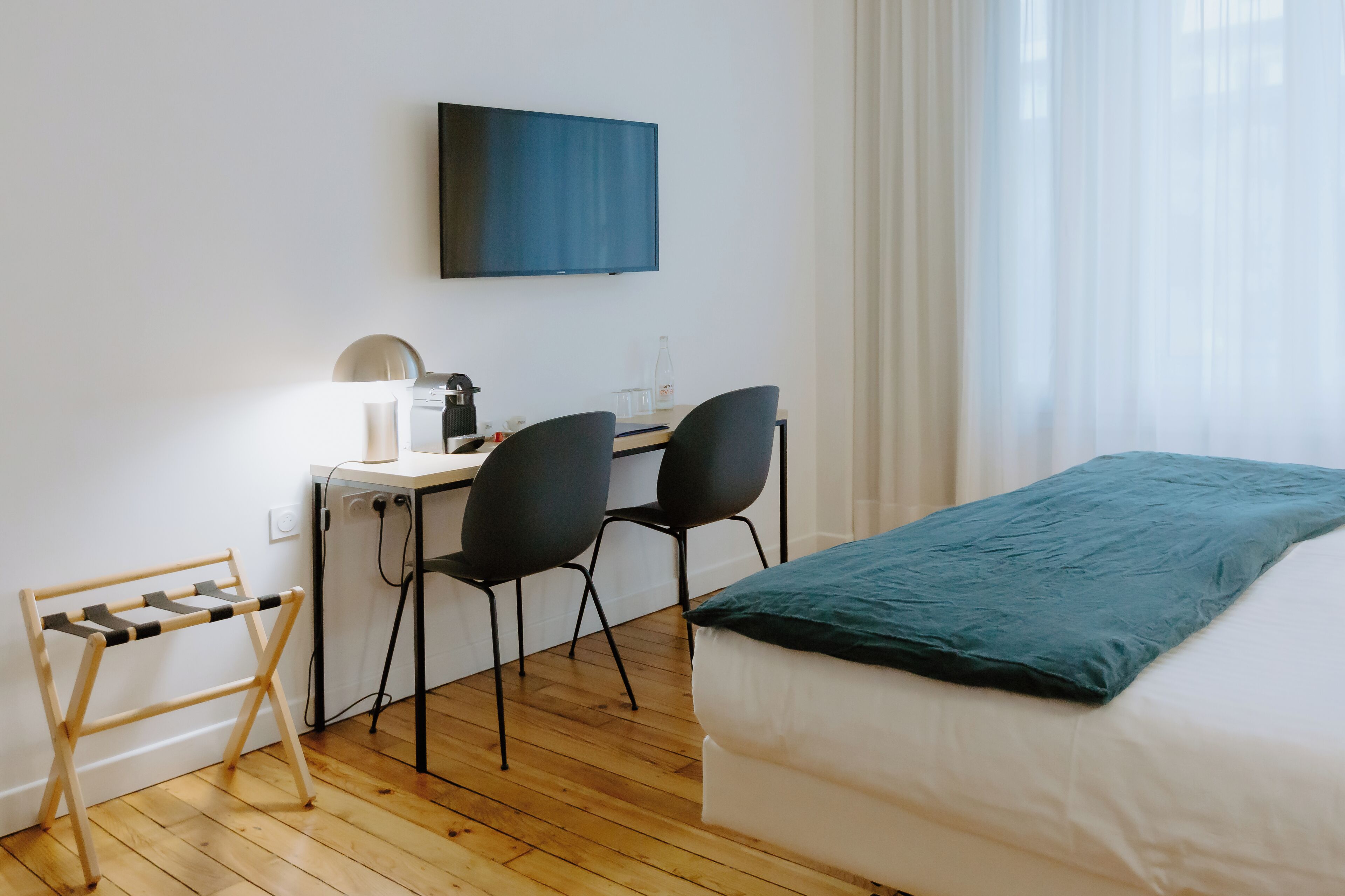 Habitación de hotel con cama, escritorio con silla, televisor montado en la pared y ventana con cortinas translúcidas.